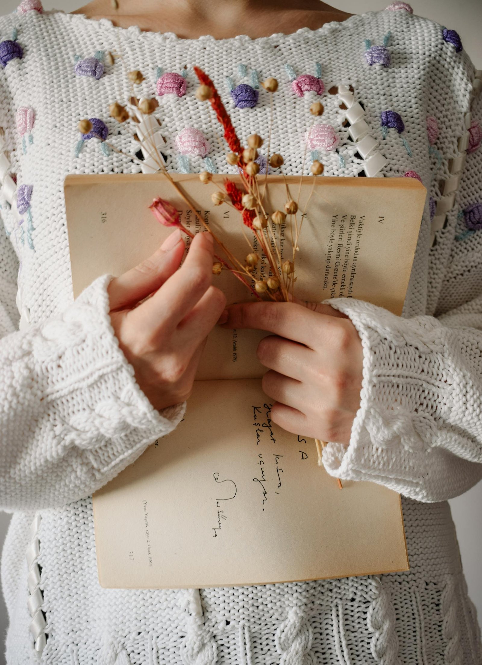 A woman in a knitted sweater holds an open book with dried flowers.