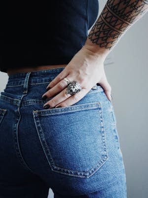 Close-up of a woman's hand with rings on her tattooed arm wearing jeans.