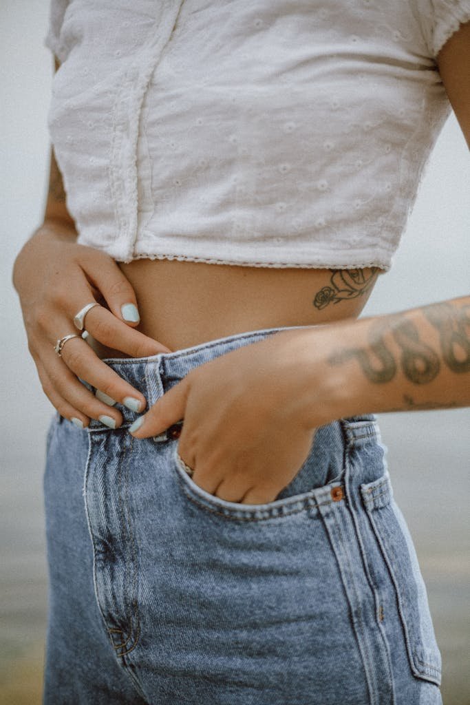 Close-up of a woman's tattooed abdomen wearing a crop top and jeans, showcasing fashion and style.