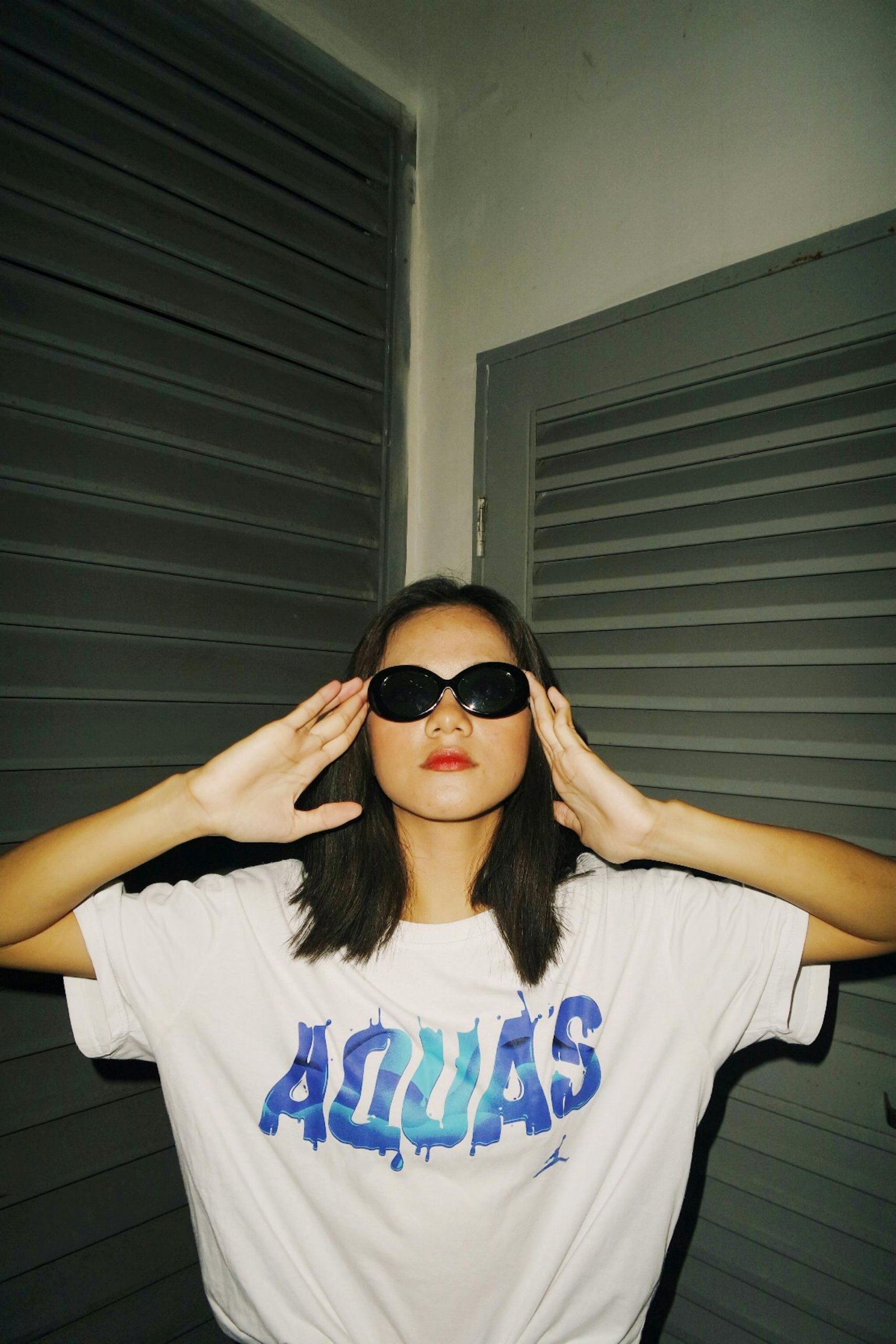 Young woman wearing sunglasses and a t-shirt, striking a pose indoors.